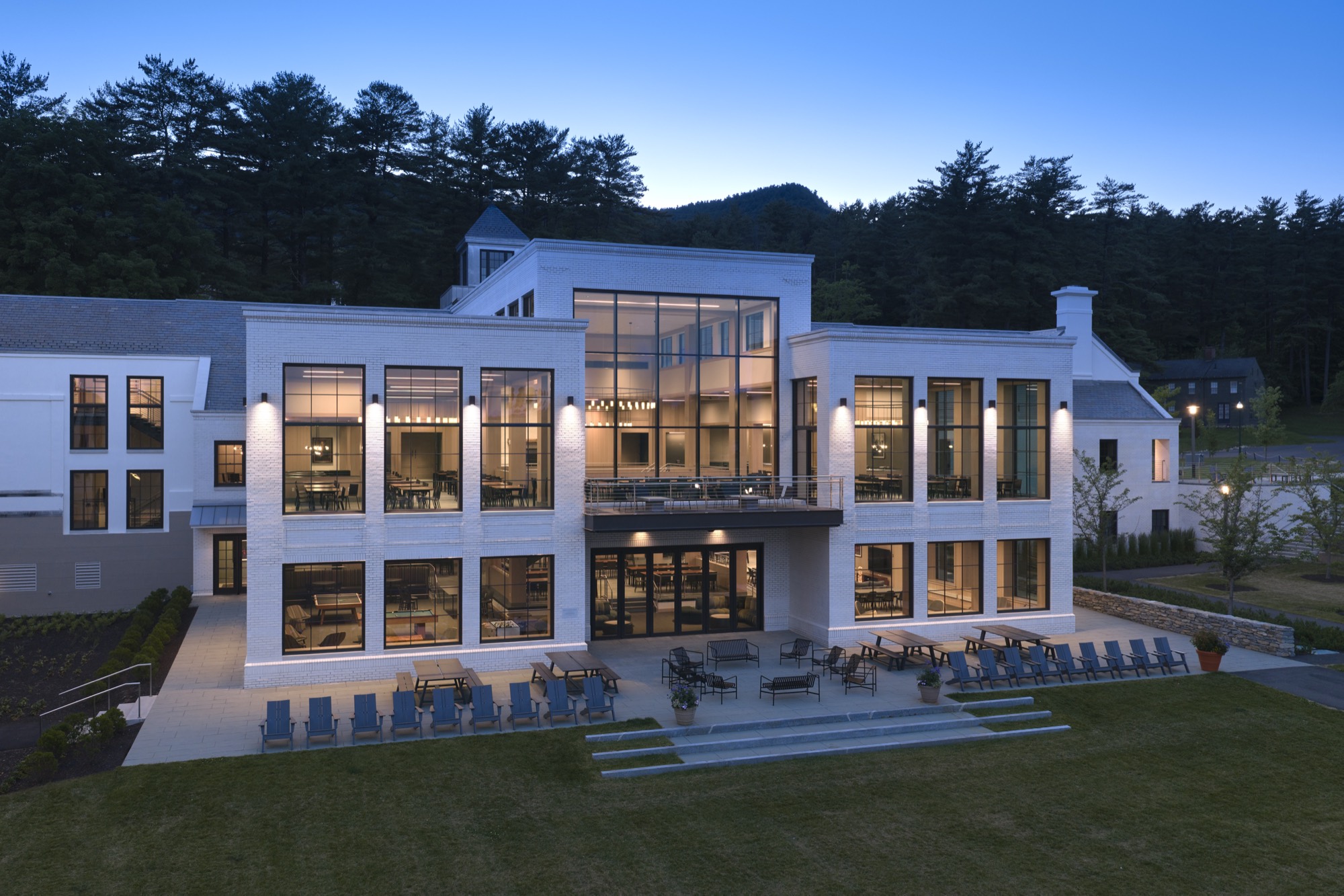Three-story white brick building with tall glass windows and a terrace at dusk